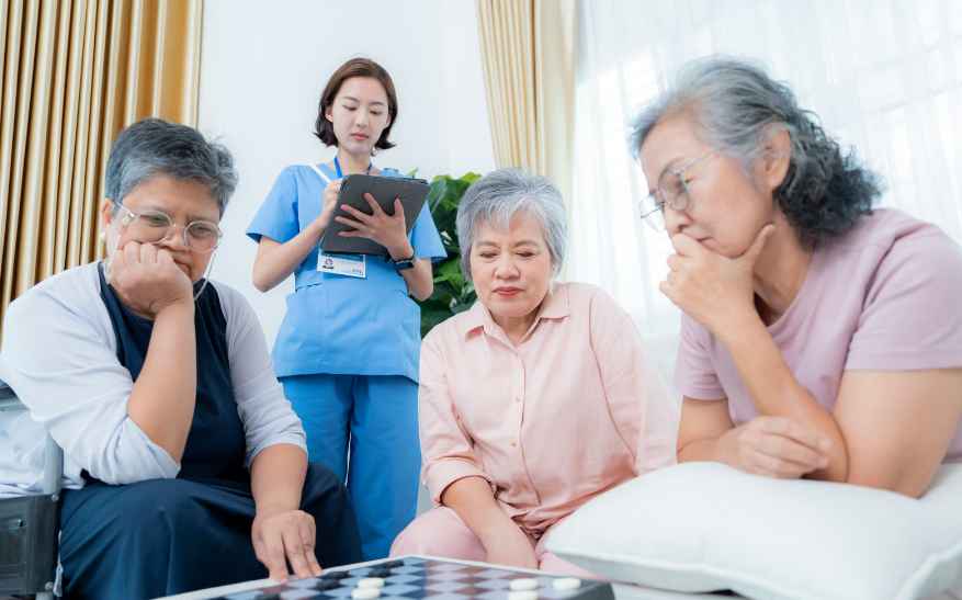 A close-up shot of three elderly Asian women deeply concentrated on a checkers match, with a friendly nurse in a blue uniform making notes on a tablet in the background.