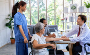 An Asian doctor holds the hand of an elderly woman in a wheelchair during a consultation, with her caregiver standing nearby and a male companion sitting behind.