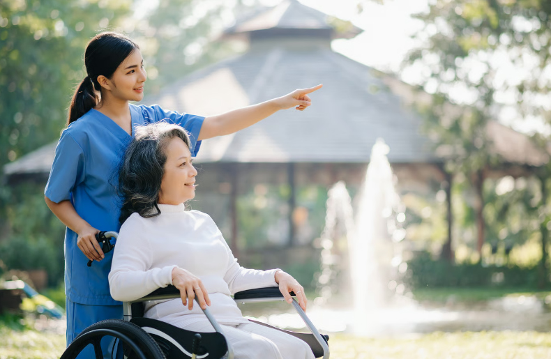 A hospice nurse in blue scrubs smiling and pointing toward a garden for a senior woman in a wheelchair.