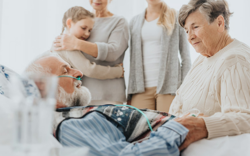 A supportive family standing by an elderly man in a hospital bed receiving palliative care.