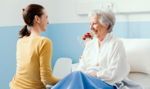 A smiling young woman in a yellow sweater sits by the hospital bed of an elderly woman with silver hair, who is laughing and looking back at her.