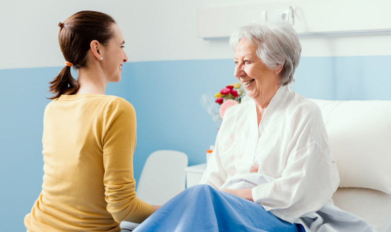 A smiling young woman in a yellow sweater sits by the hospital bed of an elderly woman with silver hair, who is laughing and looking back at her.