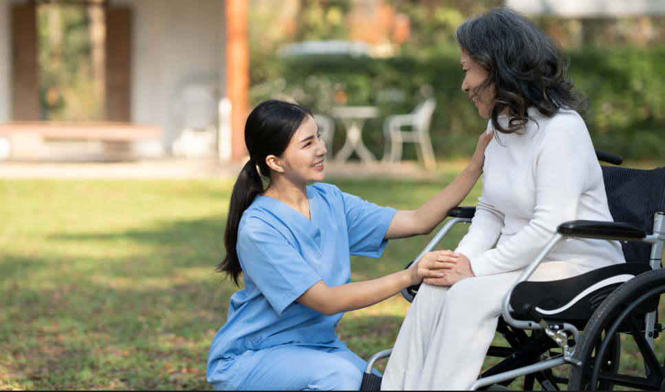 A smiling female home health nurse in blue scrubs crouches down to comfort an elderly Asian woman in a wheelchair outdoors in a garden setting.
