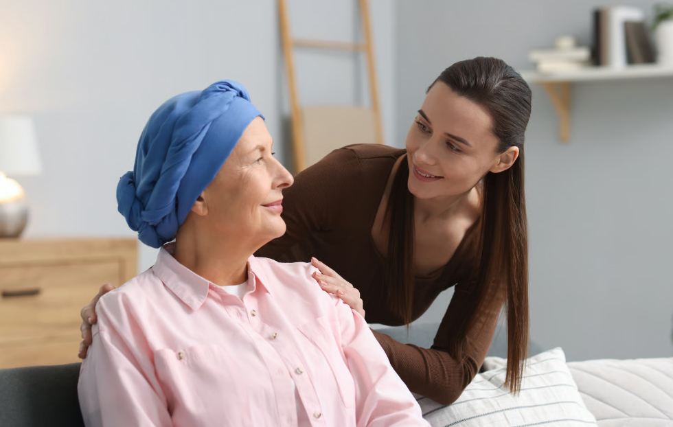 A compassionate young caregiver placing a hand on the shoulder of an elderly woman wearing a blue headscarf, illustrating the emotional support provided by a hospice care team.