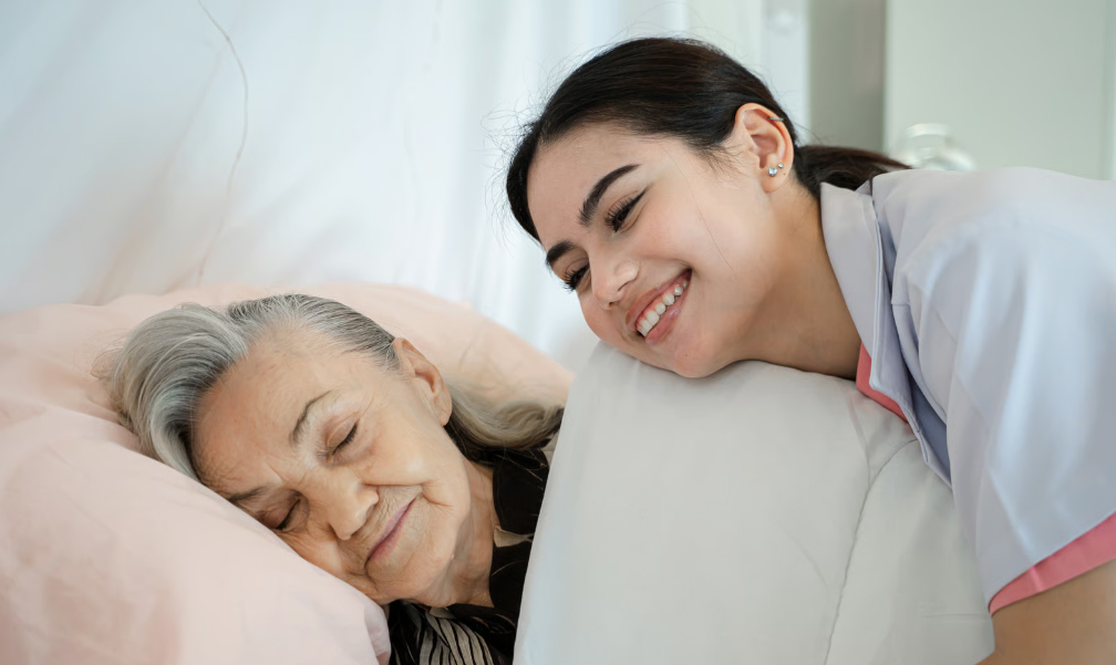 A smiling hospice nurse leaning in to care for an elderly woman resting in bed, representing the collaborative medical oversight between hospice teams and primary care physicians.