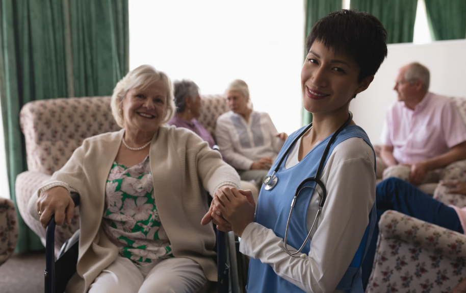 A smiling female nurse with short dark hair and a stethoscope holding hands with an elderly blonde woman in a armchair, with other senior residents socializing in a nursing home common room blurred in the background.