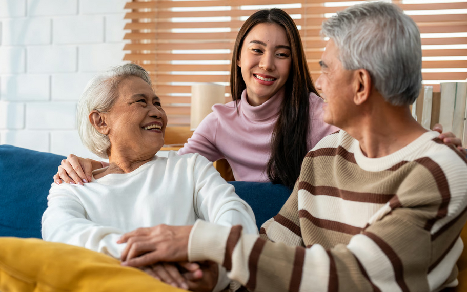 A smiling adult daughter standing behind her elderly parents on a couch, showing family comfort and caregiver relief once hospice care support begins.