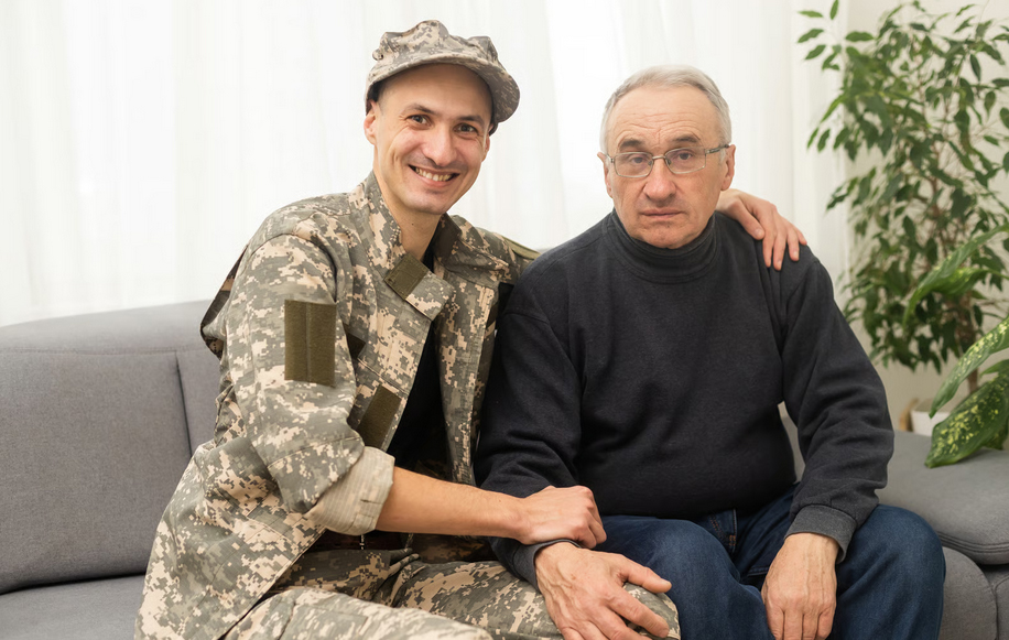 A smiling military service member in camouflage uniform sitting on a couch with an elderly male veteran with glasses, demonstrating veteran-to-veteran volunteer companionship and support during home hospice care.