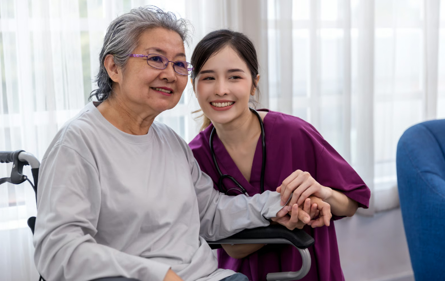 A smiling young female caregiver in purple scrubs holding the hand of an elderly Asian woman with glasses sitting comfortably in a wheelchair indoors.
