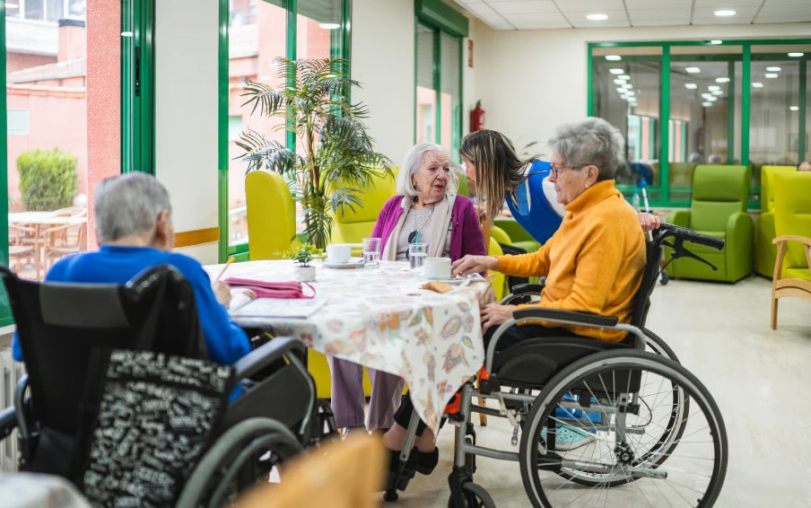 Three elderly residents, two in wheelchairs and one sitting in a chair, gathered around a dining table with coffee cups while a female staff member interacts with them in a bright common area.