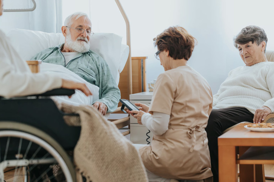 An elderly man with a white beard resting in a care bed while a nurse in a tan uniform discusses his medical plan using a device, with a family member sitting nearby.