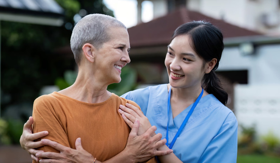 A smiling young female hospice nurse in blue medical scrubs supportively placing her hands on the shoulder of a senior woman with short gray hair outdoors, illustrating the compassionate professional support and relief families receive during scheduled respite care.