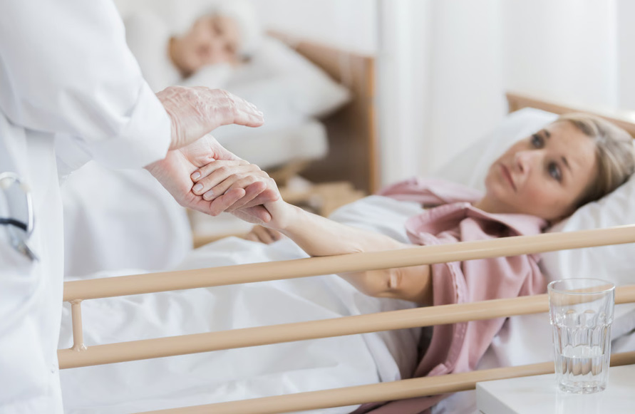 A doctor in a white coat gently holding the hand of a female cancer patient resting in a hospital bed.