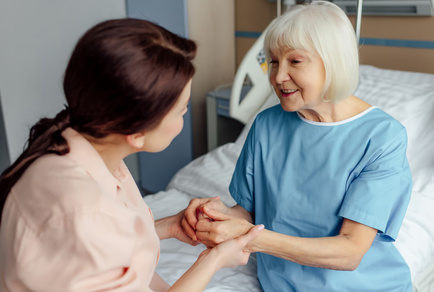 Caregiver holding hands with an elderly woman sitting on a medical bed, offering comfort and support.