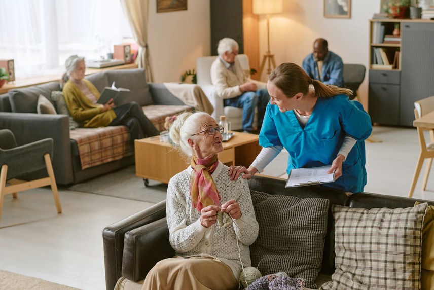 A hospice nurse in blue scrubs leaning down to speak gently with an elderly woman knitting in a living room.
