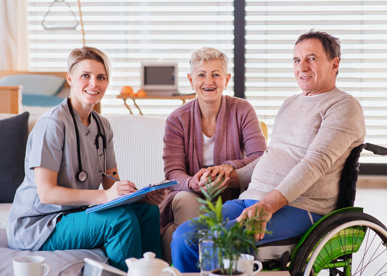 A female nurse holding a clipboard smiling with a senior male patient in a wheelchair and his wife.
