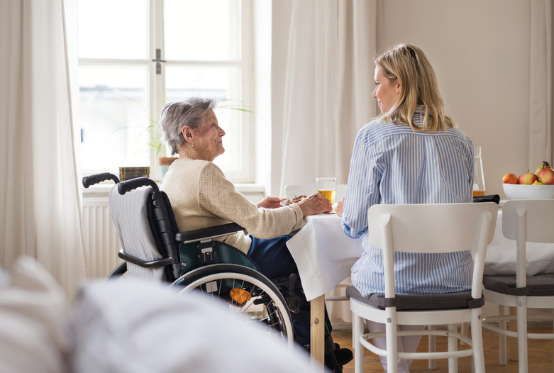A woman sitting at a dining table talking with an elderly female relative in a wheelchair near a sunlit window.